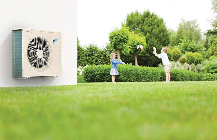 Utedel av värmepump monterad på putsad husvägg i förgrunden. Barn kastar boll i bakgrunden.